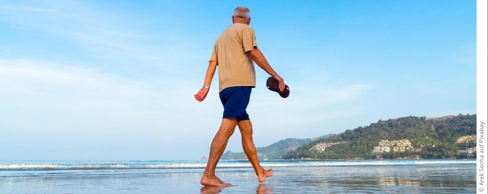 älterer Mann läuft barfuß am Strand und blickt aufs Meer